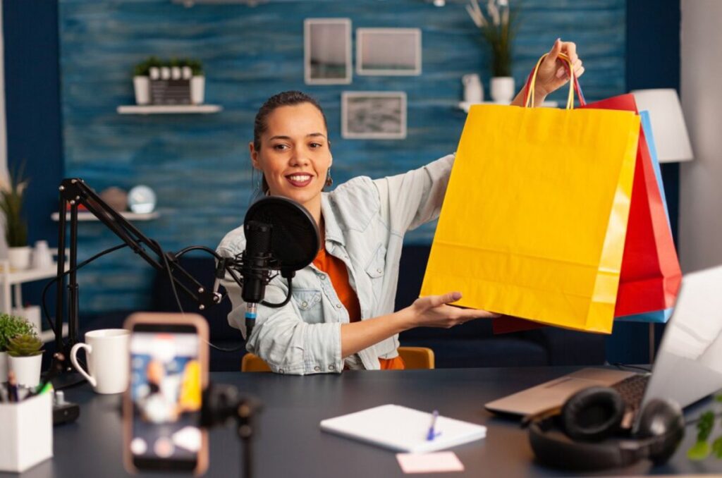 Woman in a podcast studio showcases colorful shopping bags to a microphone, with recording setup visible.