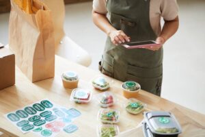 Person in apron using a tablet by table with packaged healthy meals and paper bags.