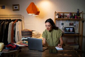 Woman working on laptop in a cozy, cluttered room with vintage decor and clothing rack.